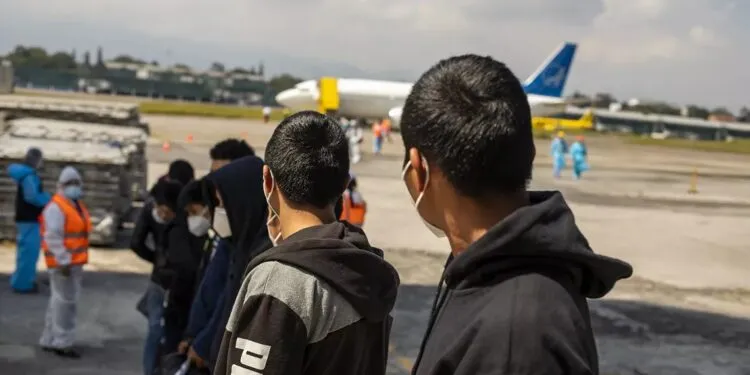 Jóvenes de espaldas con mascarillas en la pista de un aeropuerto, frente a un avión, durante un traslado relacionado con deportaciones