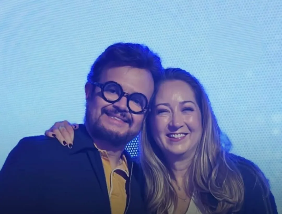 Aleks Syntek, de gafas redondas y chaqueta oscura, junto a su esposa Karen Coronado, de cabello largo rubio y sonriendo, en un evento con una pantalla azul de fondo.