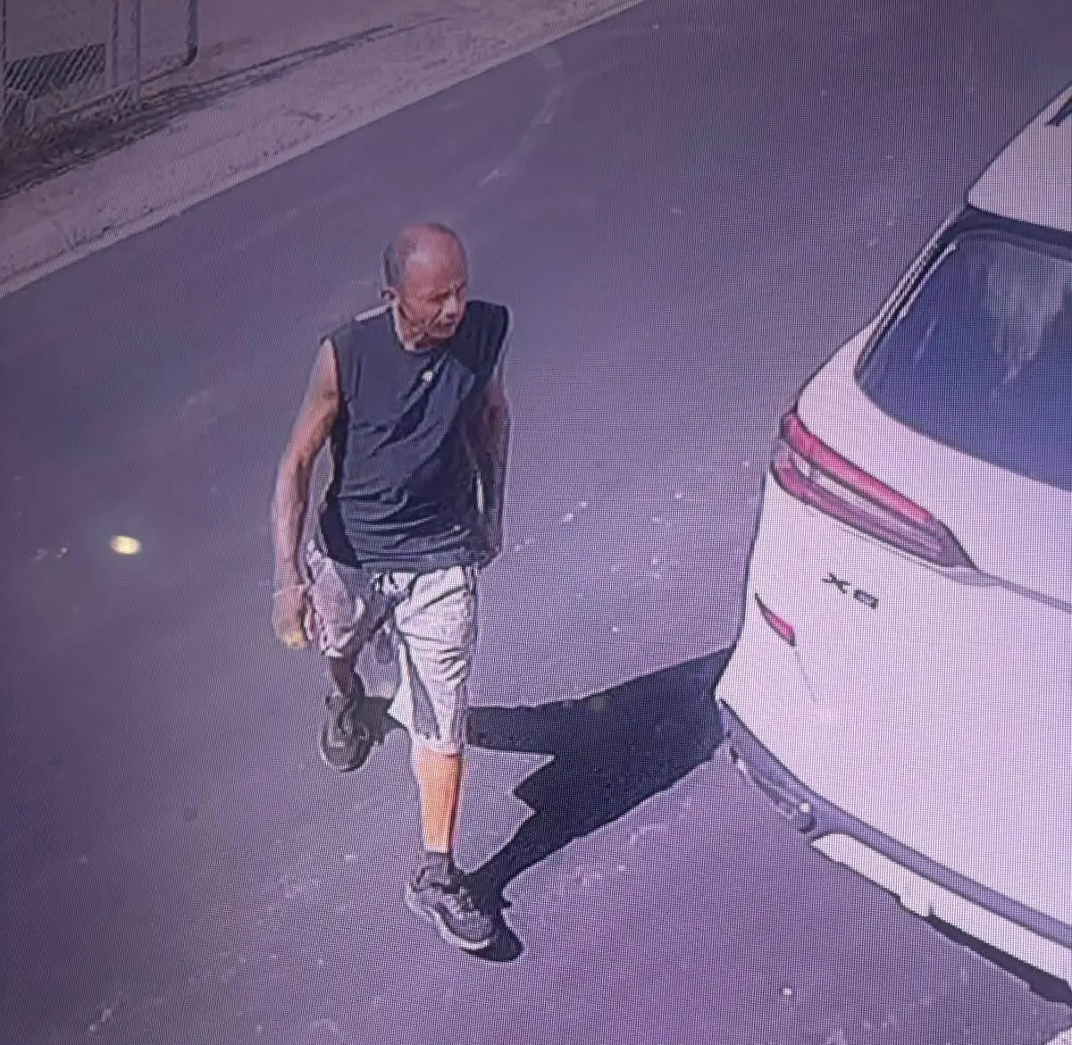 Un hombre calvo, con camiseta sin mangas negra y pantalón corto claro, camina junto a la parte trasera de una guagua blanca en un estacionamiento, captado por una cámara de seguridad.