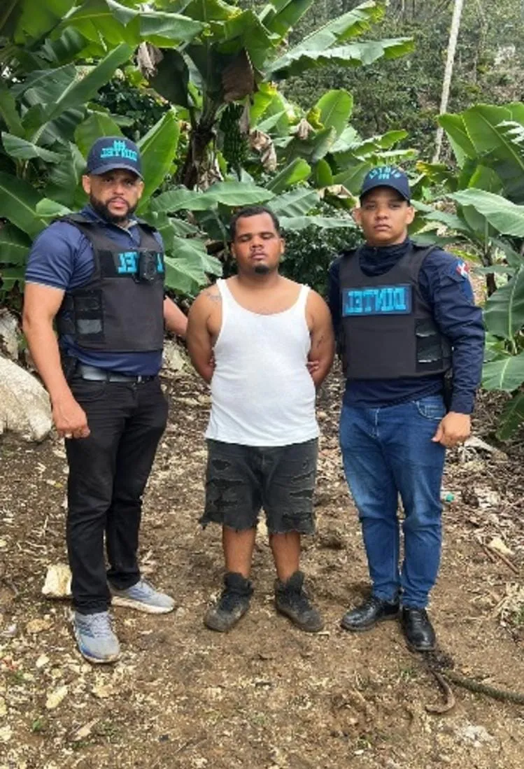 Yeisel Yoel Dotel Carvajal de pie, con las manos a la espalda, entre dos agentes del DINTEL en una zona montañosa de Bahoruco tras su captura