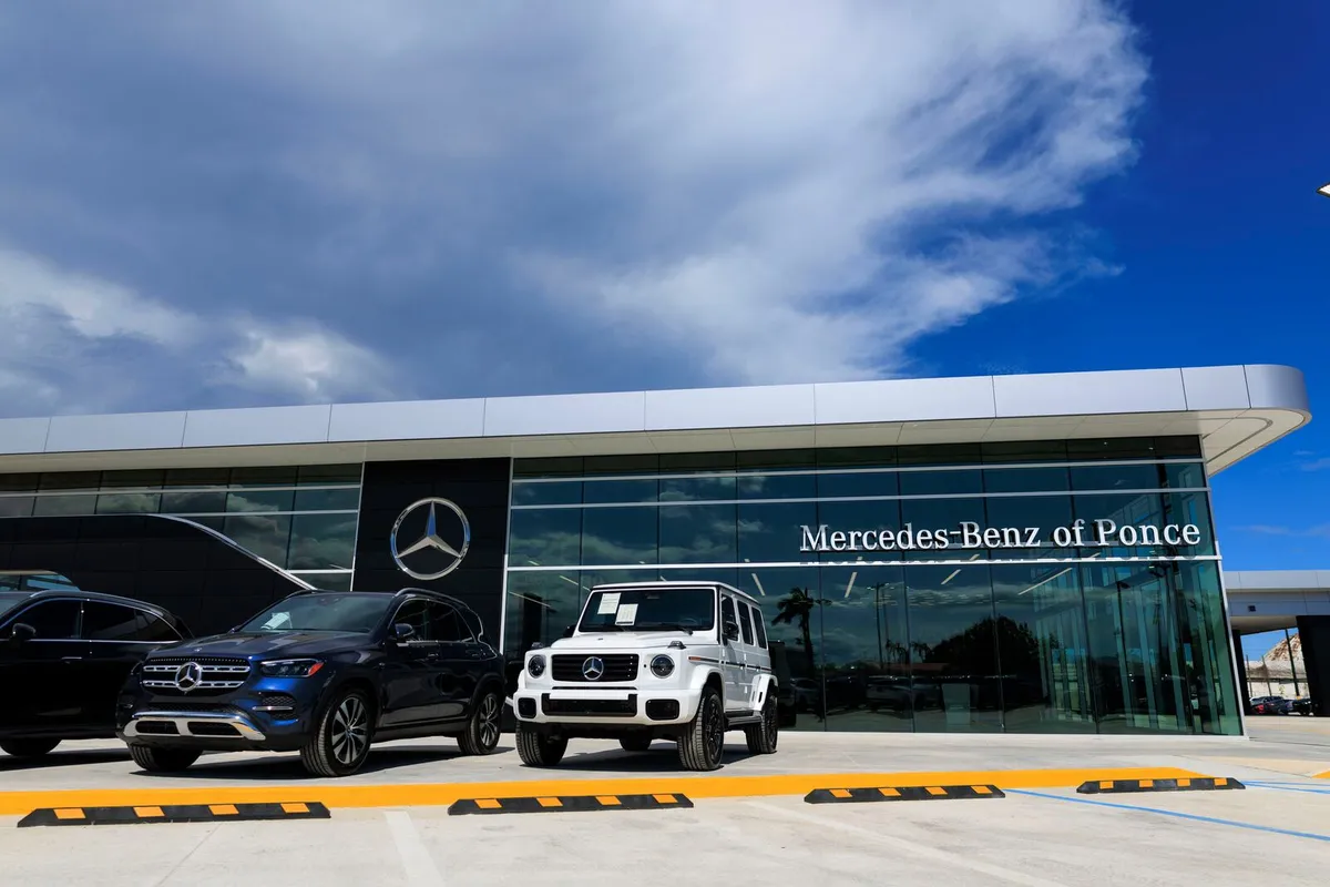 Fachada de cristal del concesionario Mercedes-Benz of Ponce bajo un cielo azul, con una guagua blanca G-Class y una SUV azul estacionadas frente al edificio
