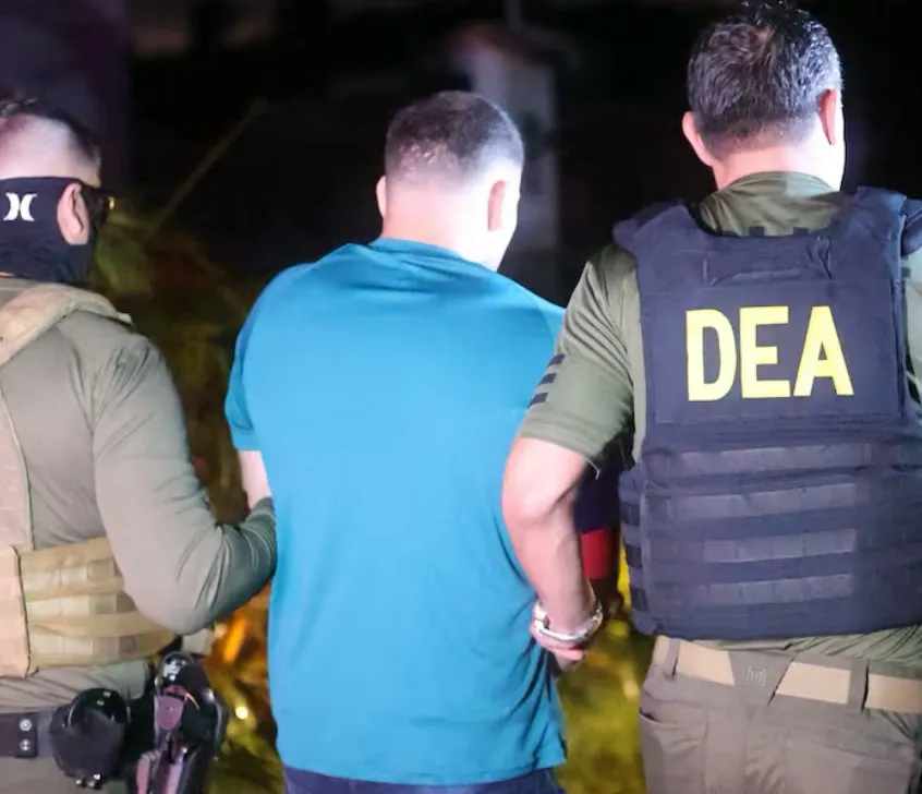 Un agente con chaleco de la DEA custodia por la espalda a uno de los detenidos.