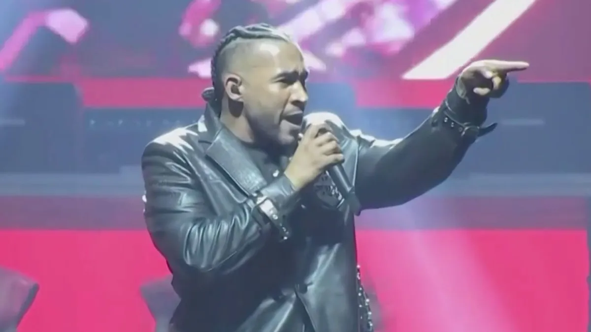 Don Omar canta con un micrófono en la mano y señala al público durante un concierto en tarima, con chaqueta de cuero negra.