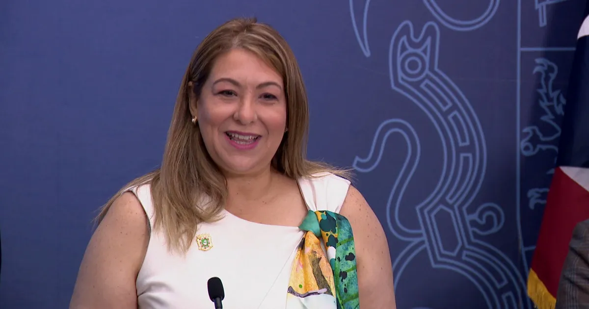 La secretaria del Departamento de la Familia, Suzanne Roig Fuertes, sonríe frente a un micrófono durante una conferencia de prensa con telón azul oficial