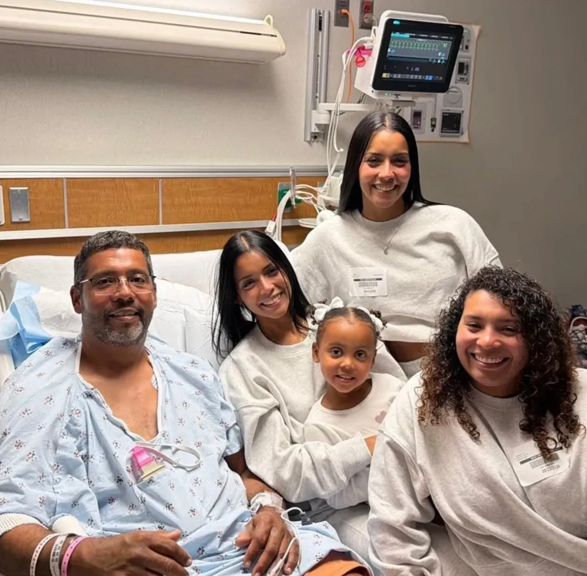 Josué Santos Rosa, con bata de hospital en una cama, sonríe junto a su esposa, dos jóvenes y una niña pequeña en un cuarto de hospital, con equipo médico al fondo.