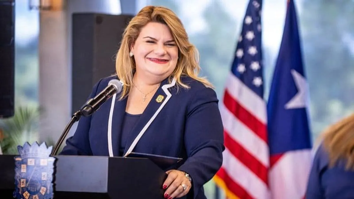 La gobernadora Jenniffer González Colón sonríe junto a un micrófono en un podio, con las banderas de Estados Unidos y Puerto Rico detrás