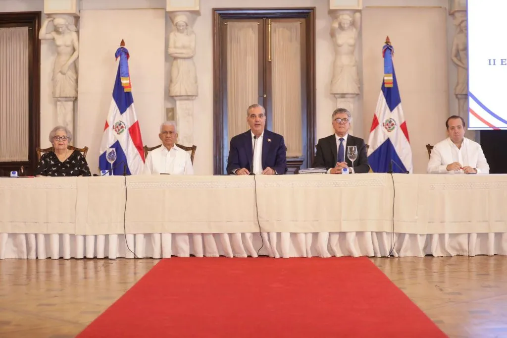 El presidente Luis Abinader, al centro de la mesa principal, junto a Roberto Santana y otros funcionarios durante el encuentro de reforma penitenciaria en el Palacio Nacional, con banderas de República Dominicana