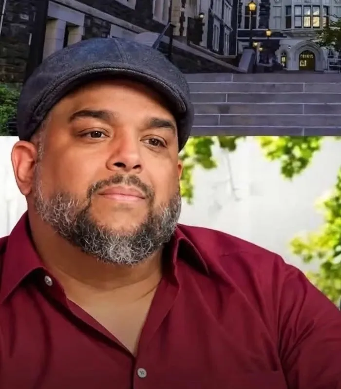 Retrato de Guillermo Rivera, de gorra gris y camisa vino, con barba canosa, frente a las escalinatas del campus del City College de Nueva York.