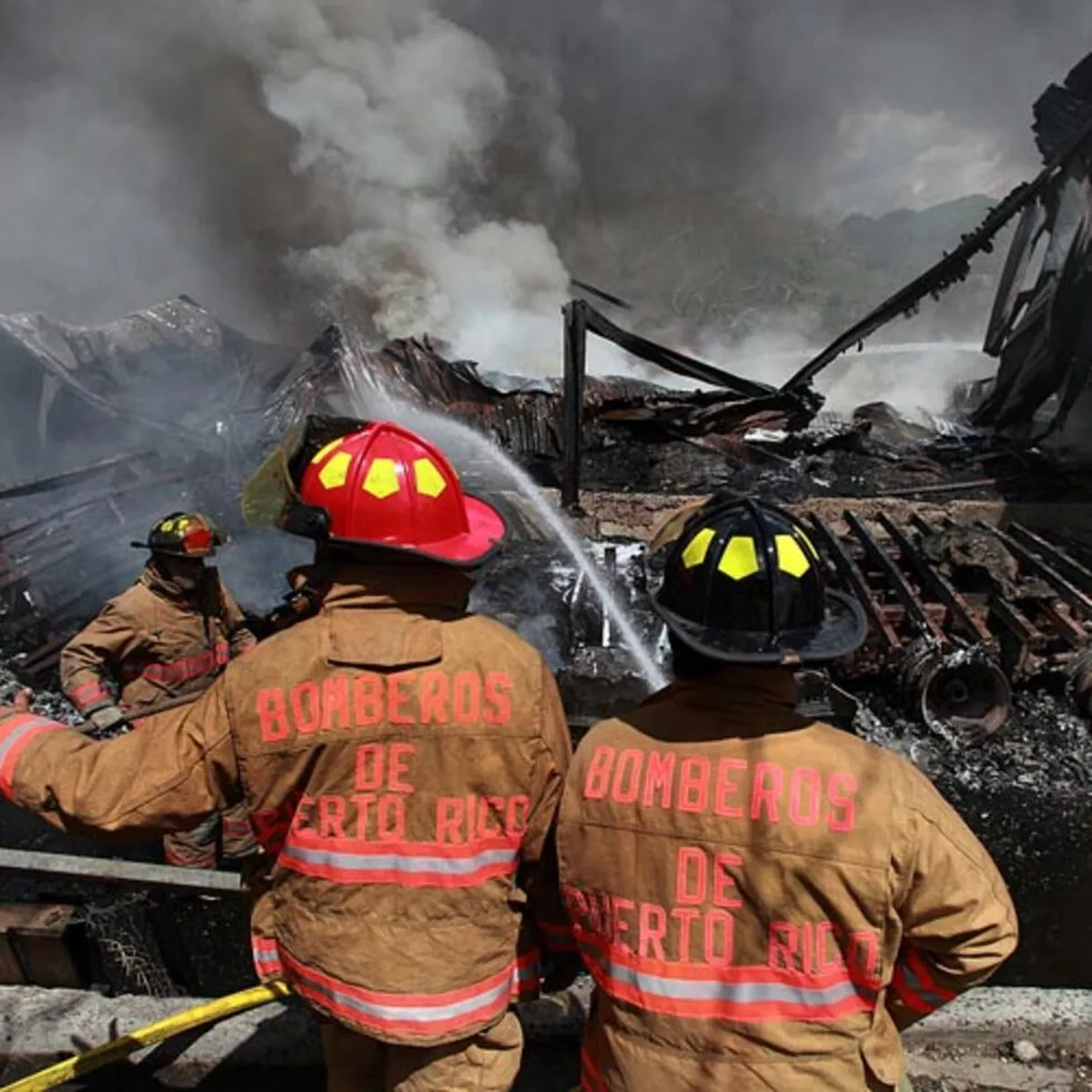 Bomberos de Puerto Rico, de espaldas, apagando con una manguera los restos humeantes de una estructura quemada, imagen de apoyo