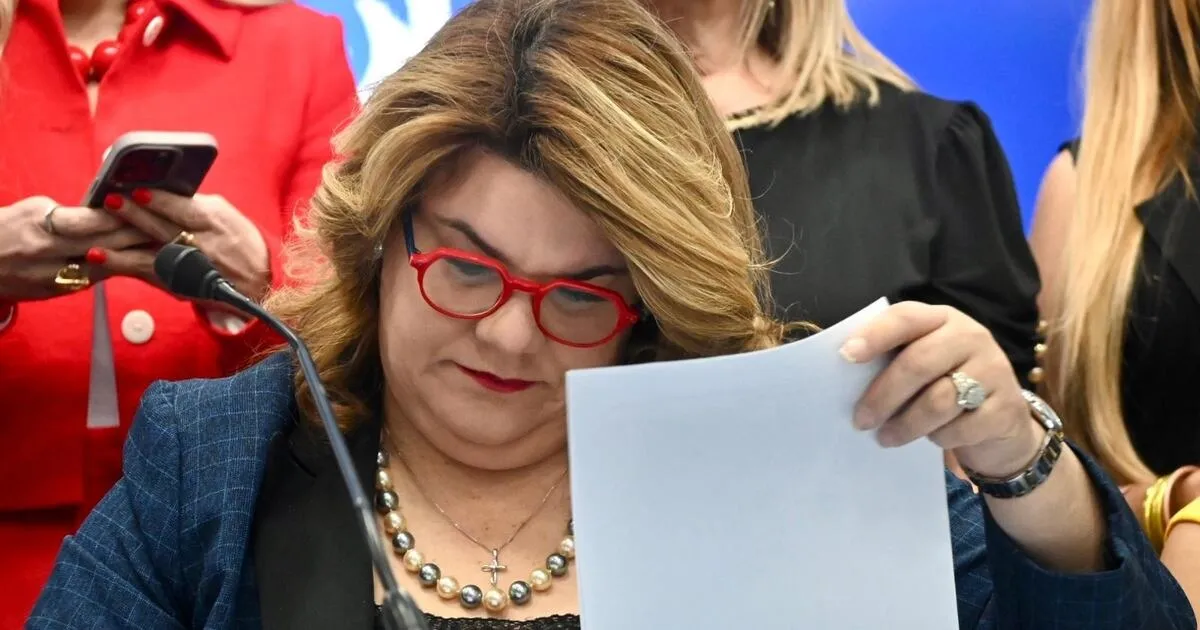 La gobernadora Jenniffer González, con gafas rojas, leyendo un documento durante una actividad oficial, rodeada de funcionarias
