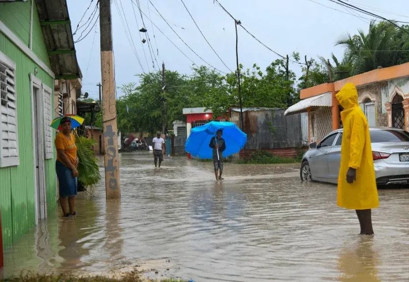Una calle anegada en una comunidad dominicana tras las lluvias, con vecinos caminando entre el agua y un joven bajo una sombrilla azul
