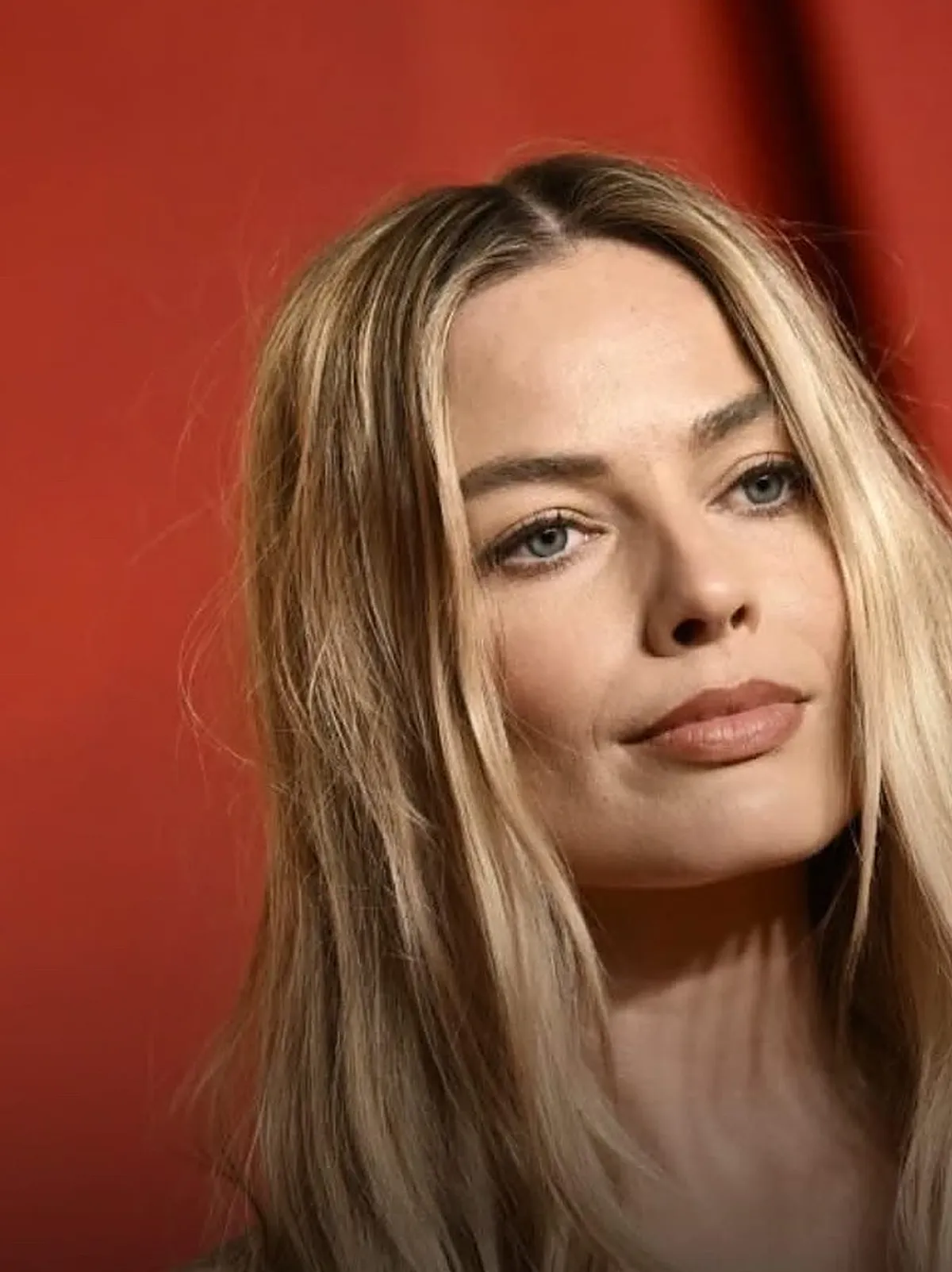 Retrato de la actriz Margot Robbie, de cabello rubio largo, sobre un fondo rojo en un evento promocional