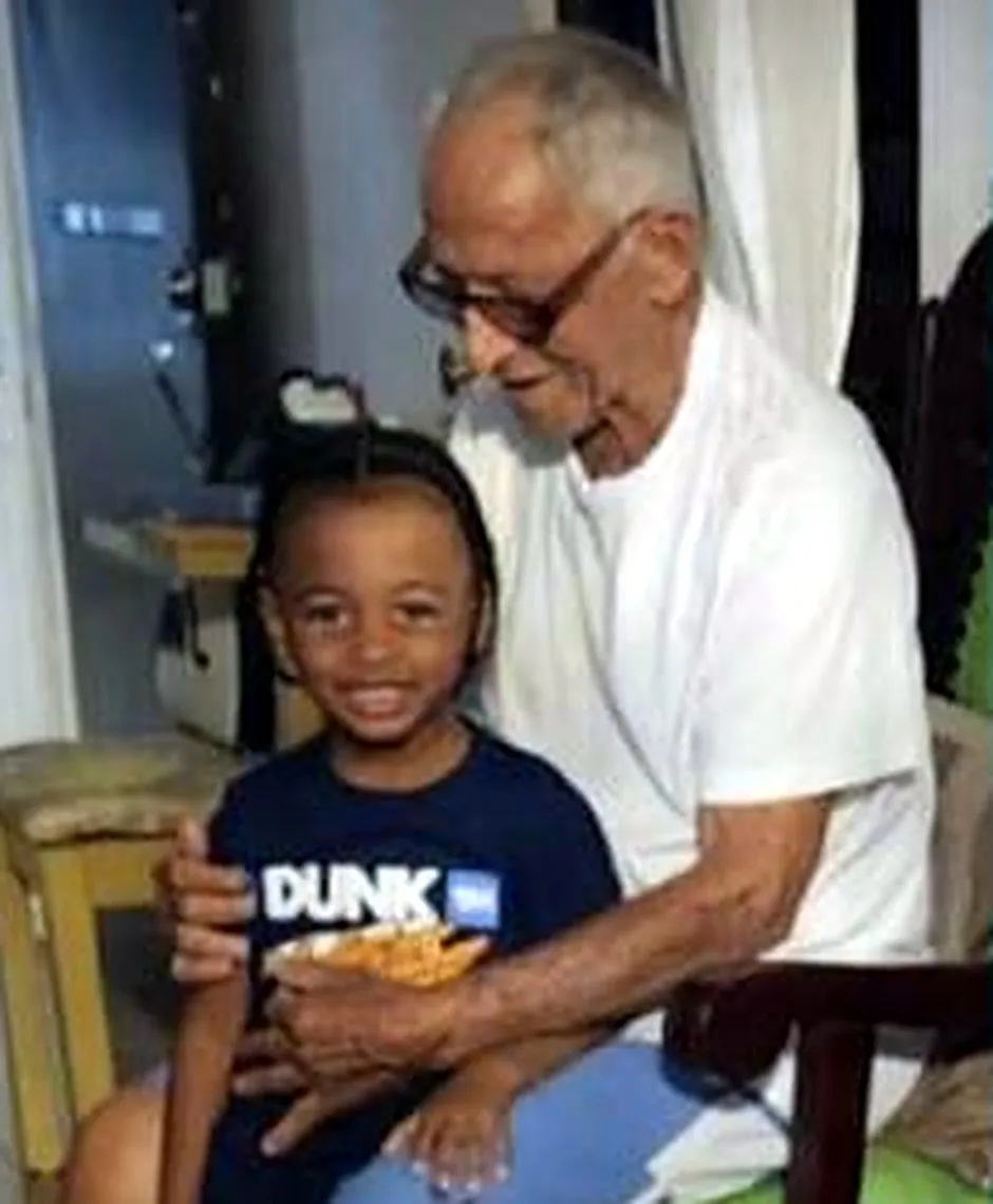 Don Santo Peña, con camiseta blanca y gafas, sentado en el interior de su casa en Castañuelas mientras abraza a su bisnieto Wave, hijo de Cardi B, que sonríe a la cámara.