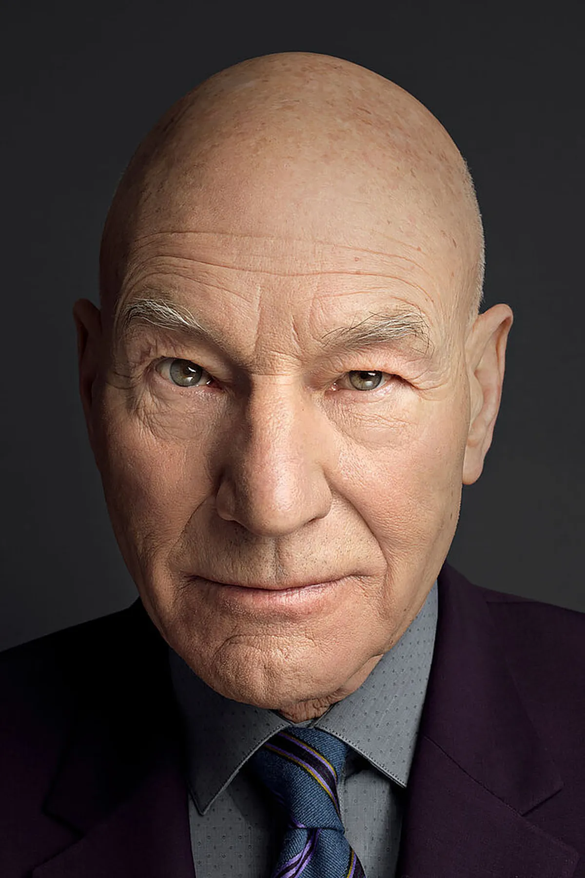 Retrato del actor británico Patrick Stewart, de traje oscuro y corbata, mirando a la cámara sobre un fondo gris oscuro