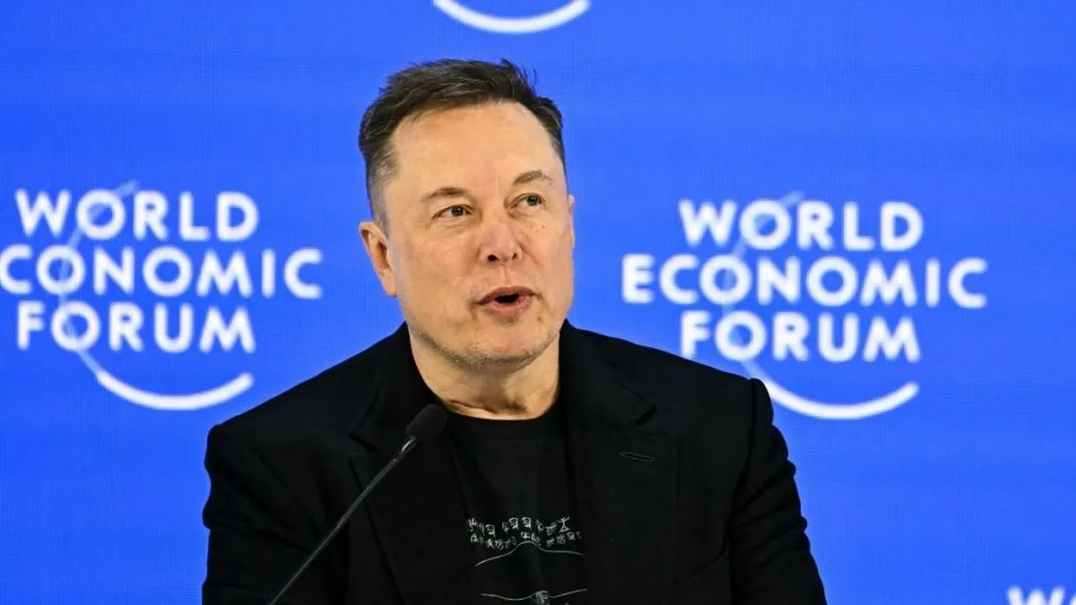 Fotografía de Elon Musk hablando ante un micrófono, con chaqueta y camiseta negras sobre un fondo azul