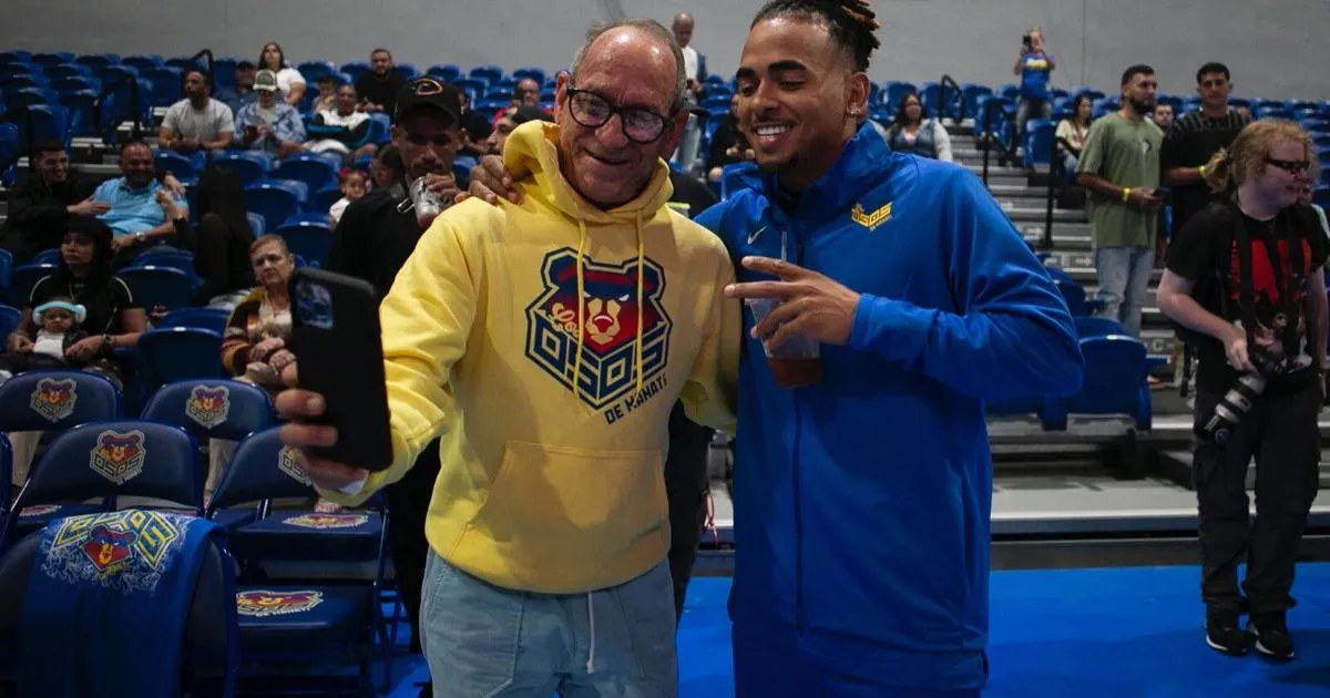 El cantante Ozuna, con una chaqueta azul de los Osos, se toma una foto con un aficionado que lleva una sudadera amarilla de los Osos de Manatí en las gradas del coliseo
