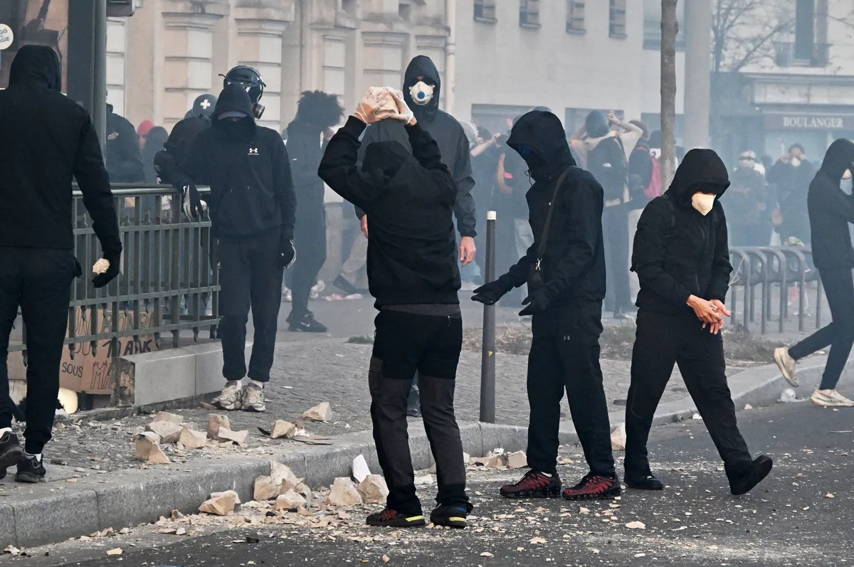 Manifestantes encapuchados entre los adoquines rotos de una calle de París, uno de ellos con una piedra levantada sobre la cabeza, durante las protestas contra el costo de la vida y la educación.