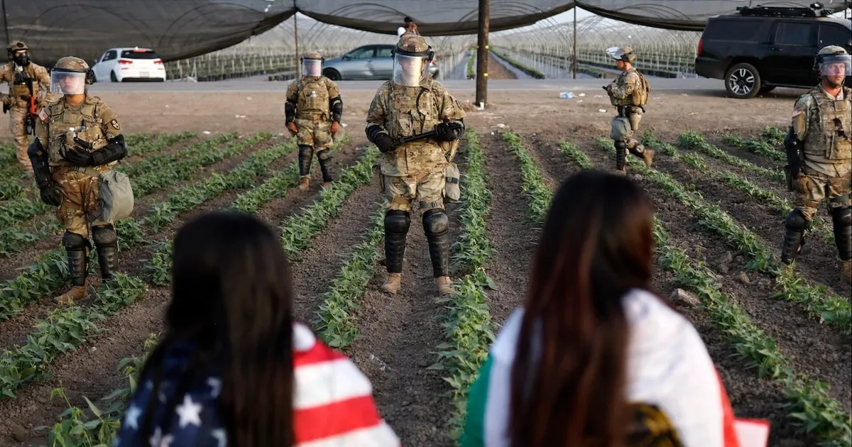 Agentes federales con equipo táctico y visores caminan entre surcos de un campo agrícola en California mientras dos mujeres los observan de espaldas en primer plano