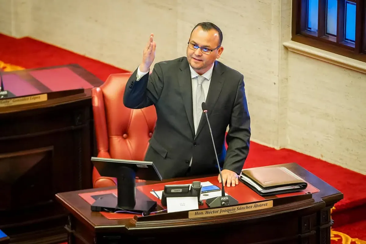 El senador Héctor Joaquín Sánchez Álvarez de pie en su escaño del hemiciclo del Senado, con la mano levantada junto al micrófono y su rótulo de nombre sobre el escritorio