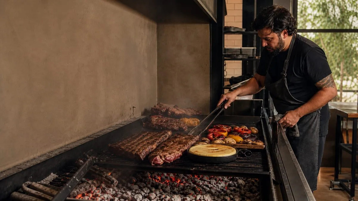 Un cocinero con delantal oscuro trabaja frente a una parrilla de carbón con carnes y vegetales en la cocina de un restaurante.