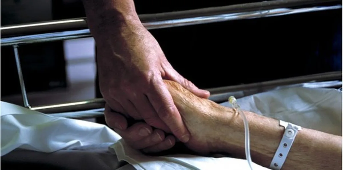 Una mano sostiene la mano de un adulto mayor acostado en una cama de hospital, con pulsera de paciente y una vía intravenosa