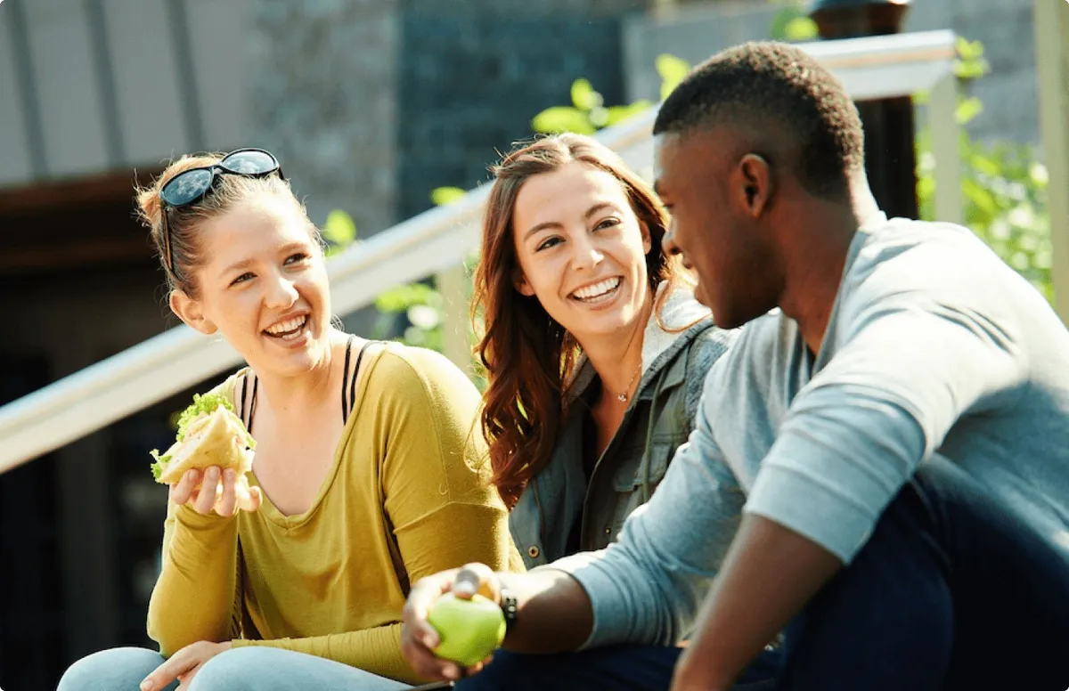 Imagen de contexto: tres jóvenes universitarios comiendo al aire libre en un campus, una de ellas con un sándwich en la mano y otro sosteniendo una manzana verde