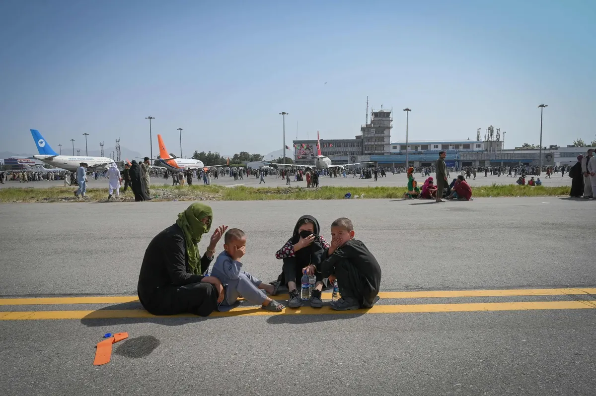 Una madre afgana con velo verde, sentada en la pista del aeropuerto de Kabul junto a tres niños, con aviones y la terminal al fondo