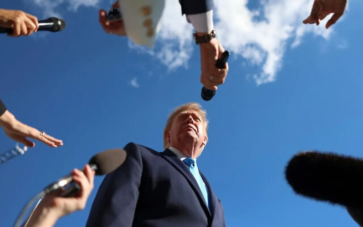 El presidente Donald Trump, rodeado de micrófonos de periodistas, hablando bajo un cielo azul con nubes