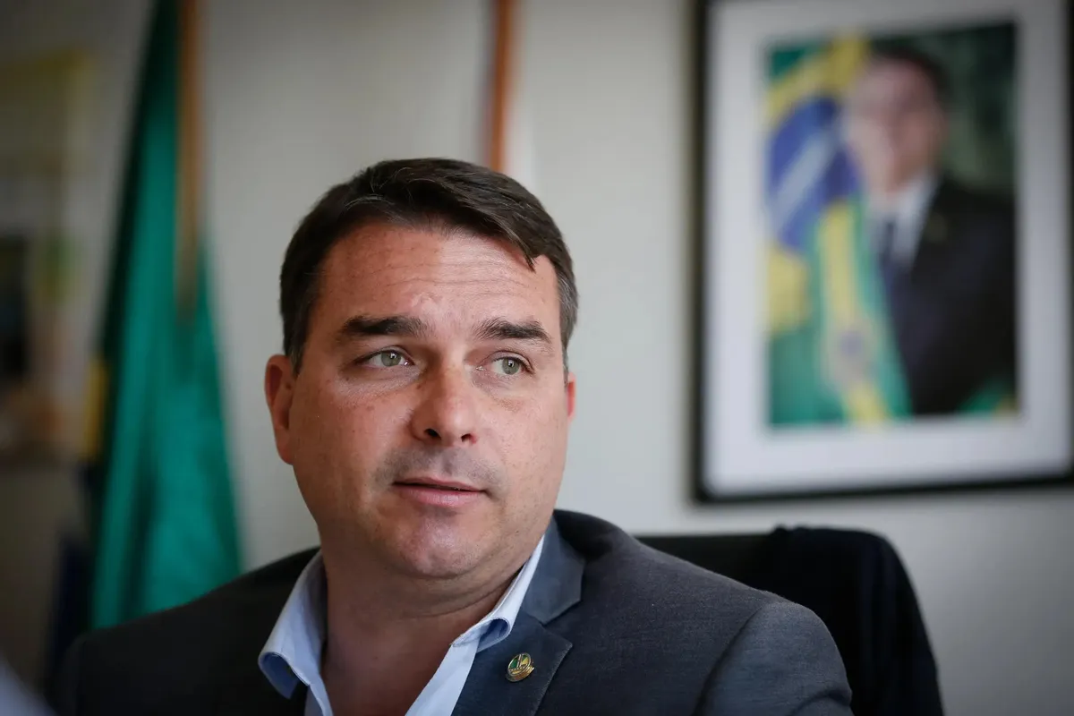Flávio Bolsonaro, en traje oscuro y sin corbata, mirando hacia un lado durante una entrevista en una oficina, con una bandera de Brasil al fondo