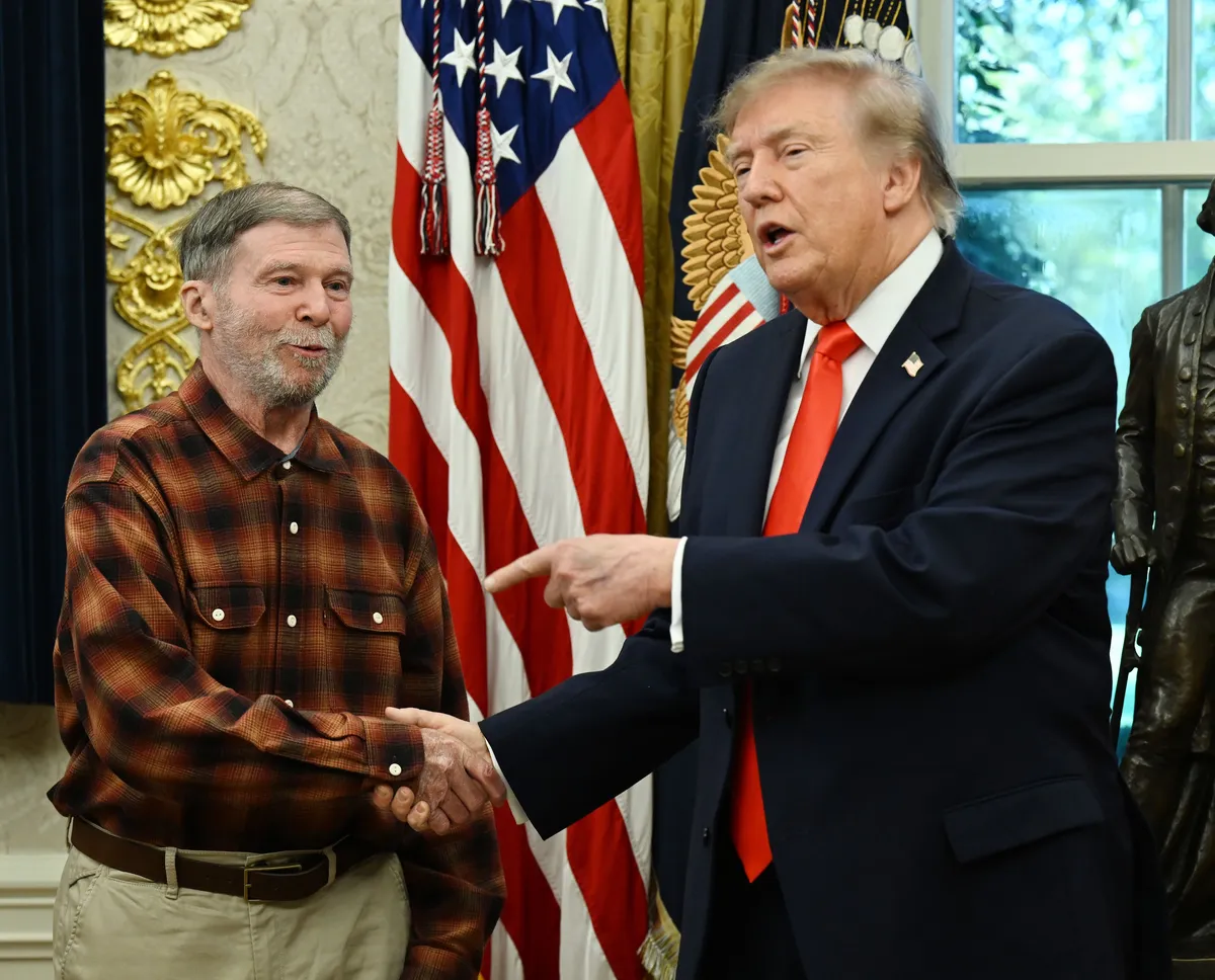 El presidente Donald Trump estrecha la mano de Stephen Hubbard, de camisa de cuadros y barba gris, en el Despacho Oval de la Casa Blanca, frente a la bandera de Estados Unidos.