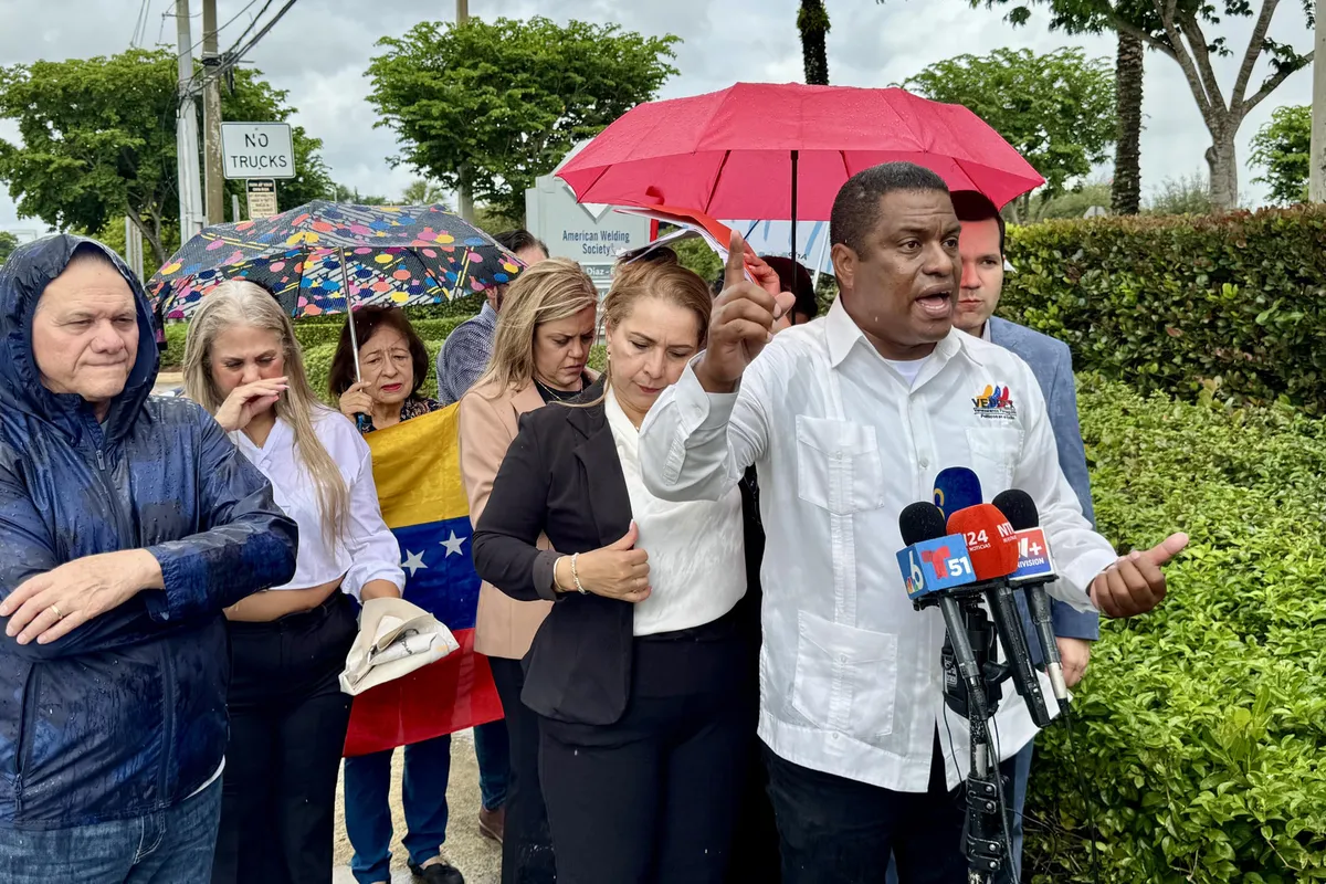 Un portavoz de VEPPEX habla ante micrófonos de prensa junto a venezolanos con una bandera de Venezuela durante una actividad en Miami